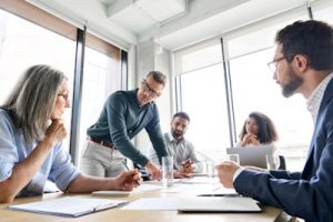 A group of professionals engaged in a strategic security discussion in a modern office. A man in business casual attire leads the conversation, emphasizing cybersecurity governance, risk management, and compliance—key aspects of Virtual CISO (vCISO) services.