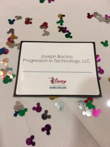 Disney Institute name badge for Joseph Bacino, Progression In Technology, LLC, displayed on a table with Mickey-shaped confetti.